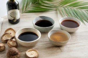 Plusieurs substituts de la sauce soja dans des bols en céramique sur bois clair : tamari, worcestershire, nuoc-mâm, aminos de coco